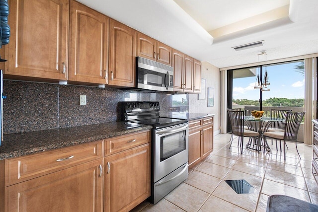 kitchen with appliances with stainless steel finishes, a tray ceiling, dark stone countertops, decorative backsplash, and light tile patterned floors