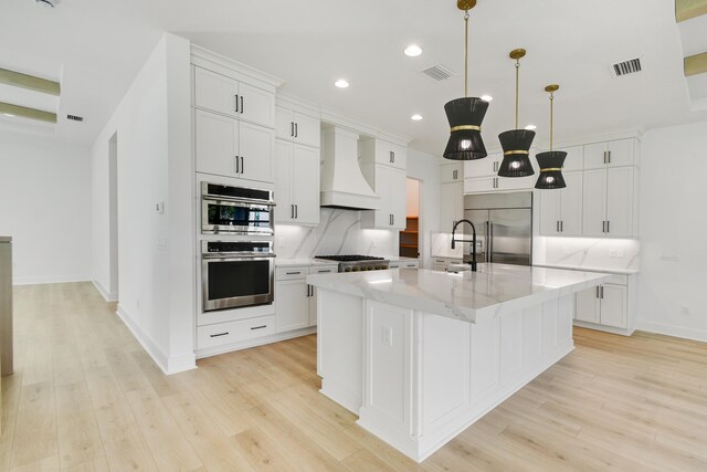 kitchen with an island with sink, custom exhaust hood, appliances with stainless steel finishes, light hardwood / wood-style floors, and tasteful backsplash