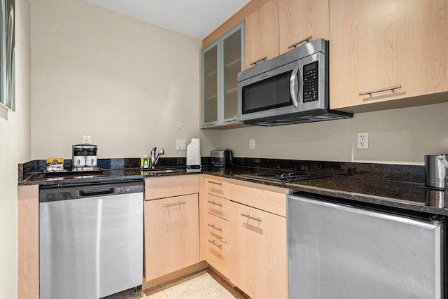 kitchen with a sink, appliances with stainless steel finishes, light brown cabinetry, dark stone countertops, and glass insert cabinets