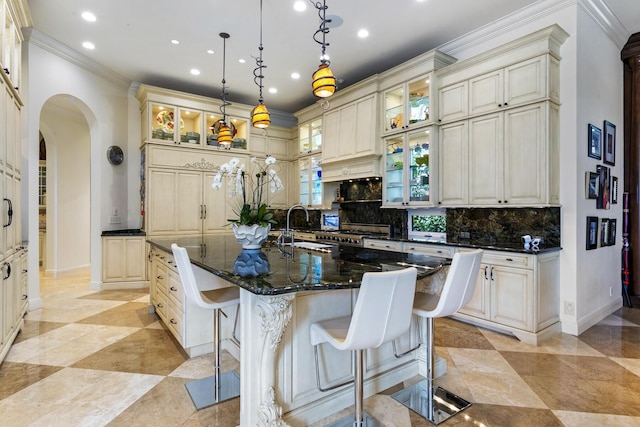 kitchen with arched walkways, a kitchen island with sink, cream cabinets, and decorative backsplash