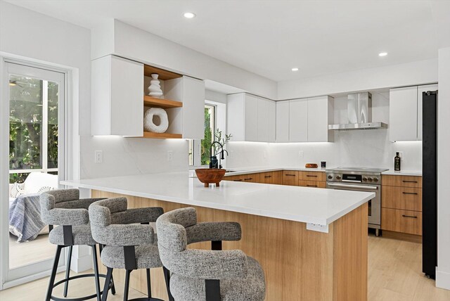 kitchen featuring a breakfast bar area, high end stainless steel range oven, a healthy amount of sunlight, wall chimney range hood, and white cabinets