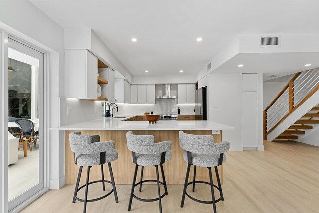 kitchen with a breakfast bar area, light wood-type flooring, wall chimney range hood, and white cabinetry