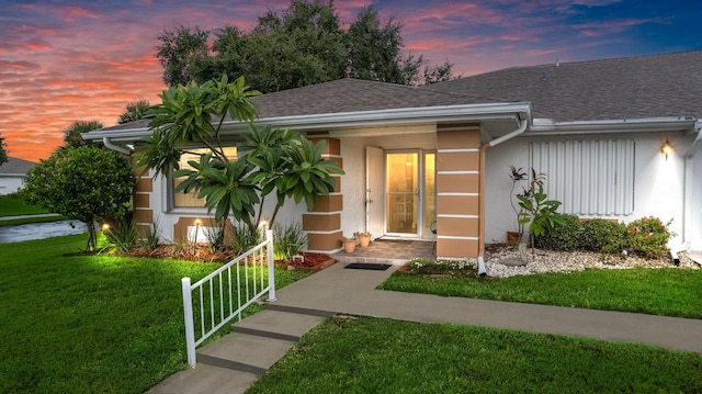 exterior entry at dusk with roof with shingles, a yard, and stucco siding