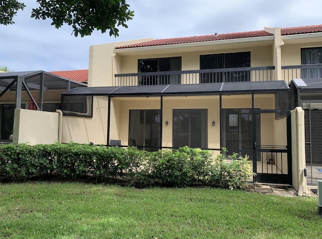 back of house featuring glass enclosure, a balcony, a tiled roof, a lawn, and stucco siding