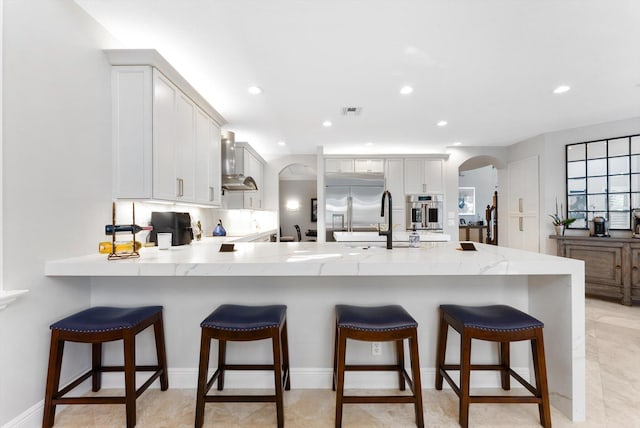 kitchen with a peninsula, arched walkways, stainless steel appliances, wall chimney range hood, and backsplash