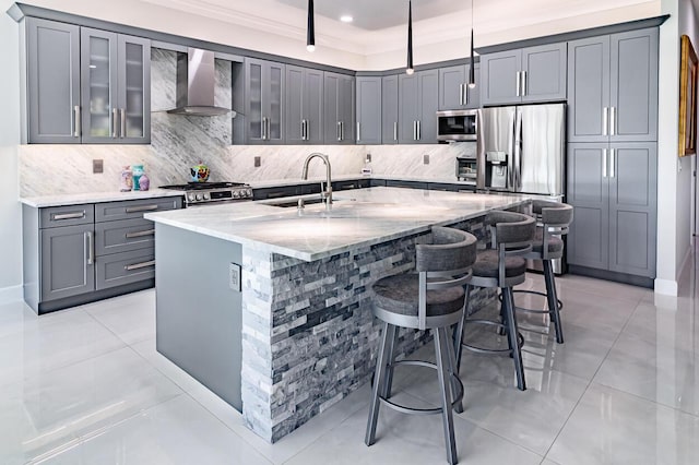 kitchen with gray cabinets, a sink, light stone counters, stainless steel appliances, and wall chimney exhaust hood