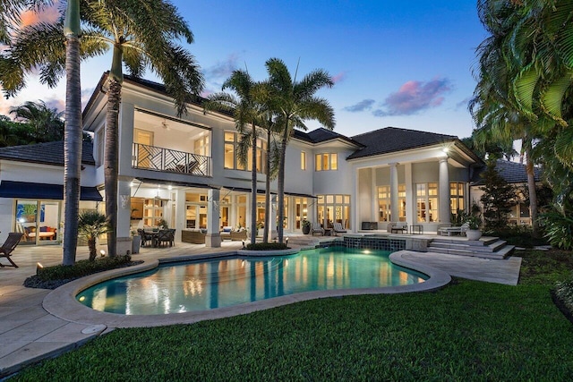 back house at dusk with a balcony, ceiling fan, a lawn, and a patio area