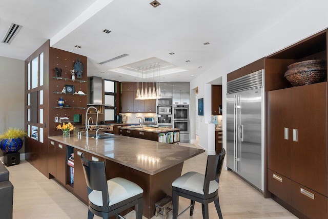 kitchen featuring light wood-type flooring, stainless steel appliances, an island with sink, hanging light fixtures, and a raised ceiling