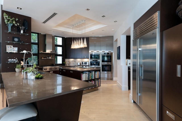 kitchen with built in appliances, pendant lighting, light hardwood / wood-style flooring, a tray ceiling, and a kitchen island with sink