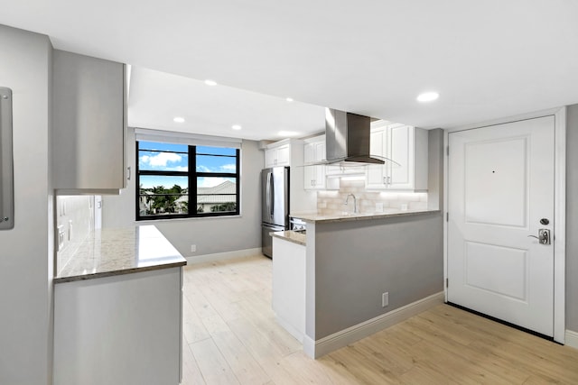 kitchen with kitchen peninsula, wall chimney exhaust hood, white cabinets, stainless steel fridge, and light wood-type flooring