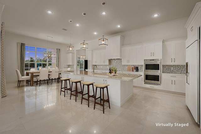 kitchen with double oven, an island with sink, white cabinets, hanging light fixtures, and light stone countertops