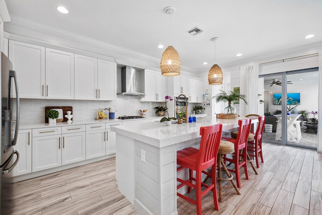 kitchen featuring a center island with sink, wall chimney range hood, hanging light fixtures, white cabinetry, and stainless steel appliances