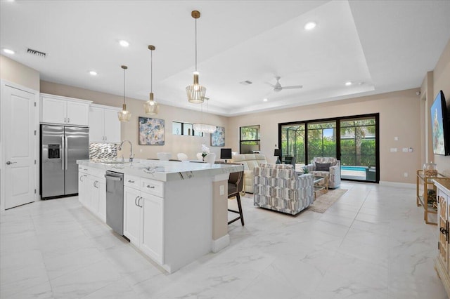 kitchen with light stone counters, a kitchen island with sink, white cabinets, stainless steel appliances, and a raised ceiling