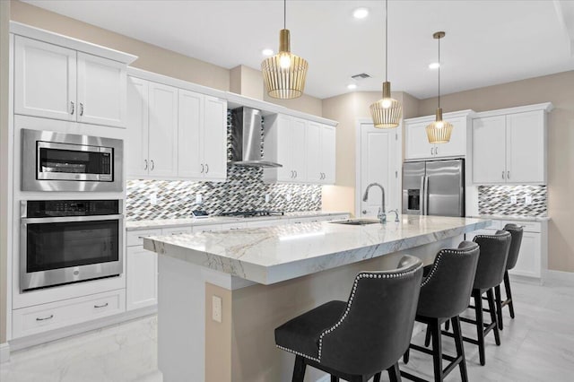 kitchen with decorative light fixtures, a kitchen island with sink, wall chimney range hood, white cabinetry, and stainless steel appliances