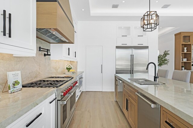 kitchen with an inviting chandelier, premium range hood, high quality appliances, white cabinetry, and light wood-type flooring