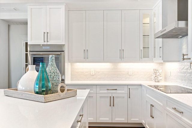 kitchen with oven, backsplash, black electric stovetop, wall chimney range hood, and white cabinets