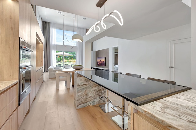 kitchen featuring light wood-type flooring, a kitchen bar, decorative light fixtures, double oven, and light brown cabinets