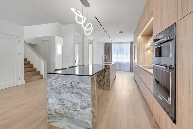 kitchen with hanging light fixtures, stainless steel double oven, light hardwood / wood-style floors, an island with sink, and black electric stovetop