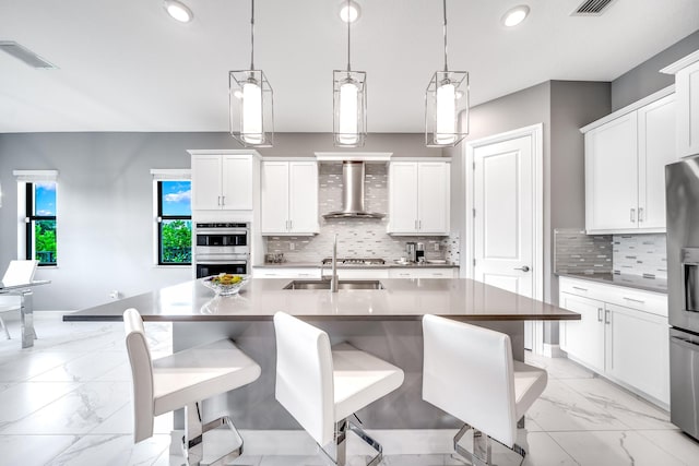 kitchen with wall chimney exhaust hood, backsplash, a center island with sink, and white cabinets