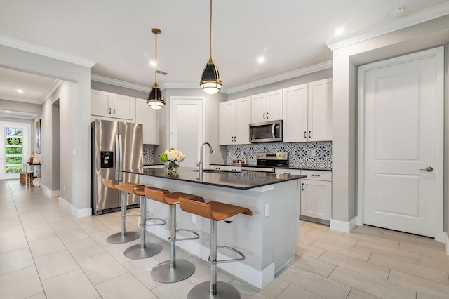 kitchen with crown molding, appliances with stainless steel finishes, white cabinetry, pendant lighting, and a center island with sink