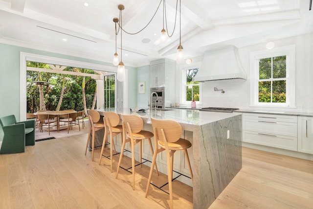 kitchen featuring custom range hood, white cabinetry, an island with sink, light stone counters, and light wood-type flooring