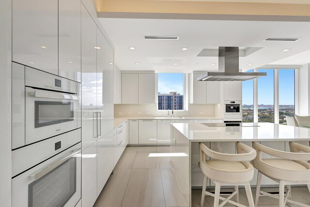 kitchen featuring a kitchen breakfast bar, island exhaust hood, decorative backsplash, sink, and white cabinetry