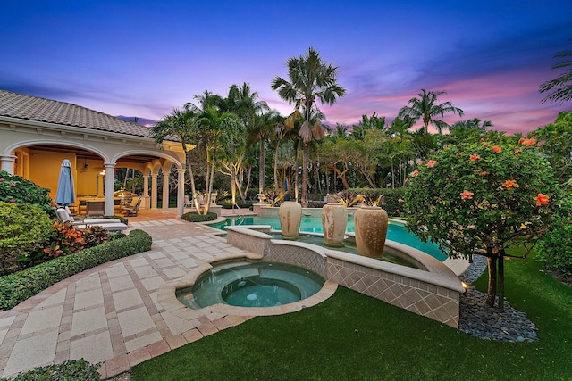 pool at dusk featuring a patio, a lawn, and an in ground hot tub