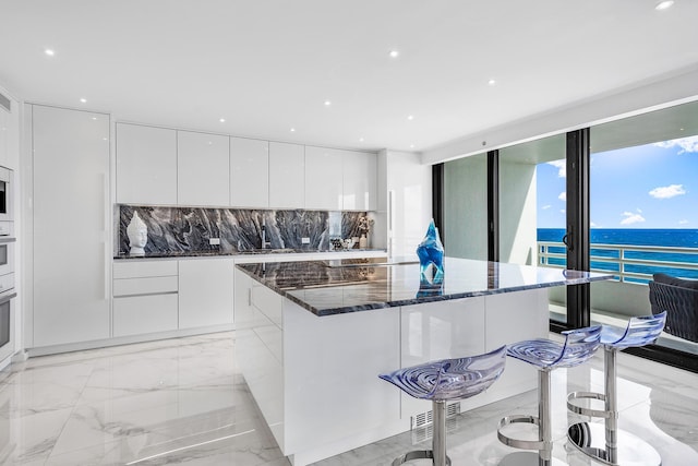 kitchen with a kitchen bar, a water view, white cabinetry, dark stone countertops, and decorative backsplash