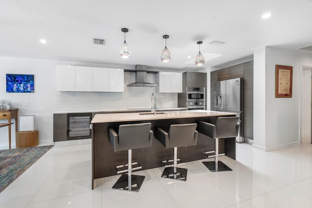 kitchen featuring decorative light fixtures, wall chimney exhaust hood, an island with sink, and light tile patterned flooring