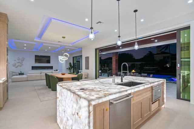 kitchen with light brown cabinetry, decorative light fixtures, stainless steel appliances, sink, and a center island with sink