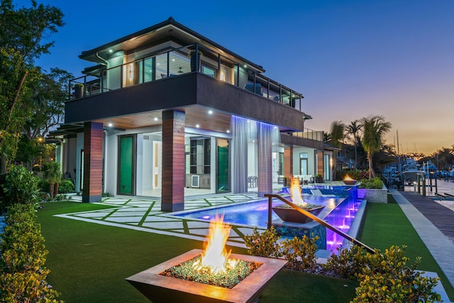 back of house at dusk featuring a patio area, a balcony, a fire pit, and an infinity pool