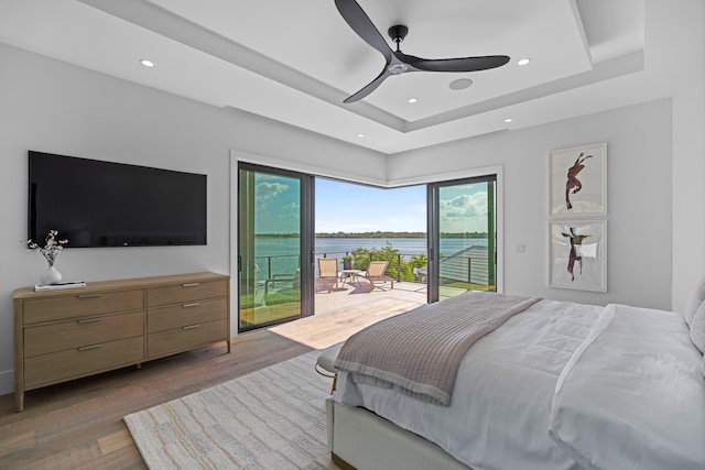 bedroom featuring a water view, light hardwood / wood-style flooring, access to exterior, ceiling fan, and a raised ceiling