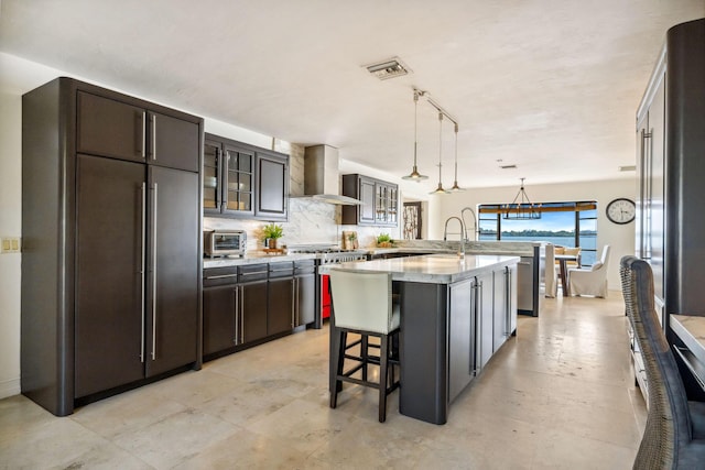 kitchen with high end appliances, light countertops, glass insert cabinets, an island with sink, and wall chimney exhaust hood