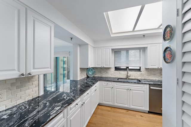 kitchen with dark stone counters, a skylight, sink, stainless steel dishwasher, and light hardwood / wood-style floors