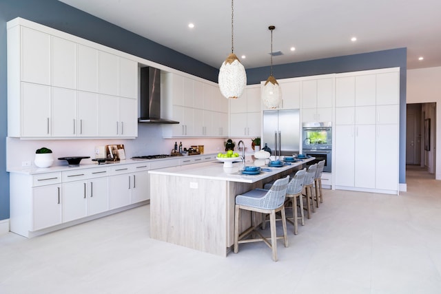 kitchen with white cabinets, stainless steel appliances, a center island with sink, and wall chimney range hood