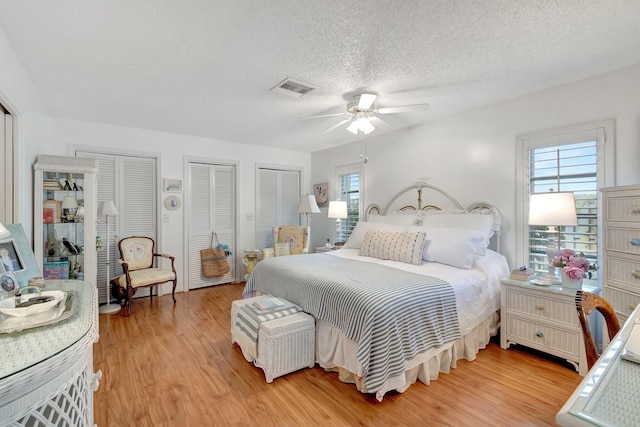 bedroom with a textured ceiling, ceiling fan, multiple closets, and light hardwood / wood-style flooring