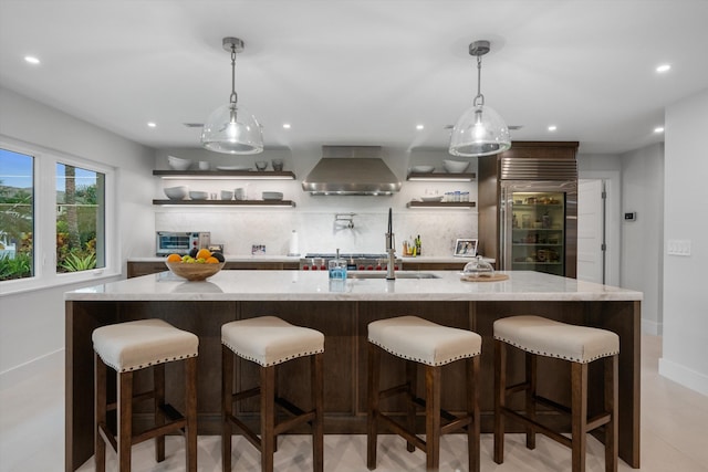 kitchen with stainless steel built in refrigerator, pendant lighting, an island with sink, a kitchen bar, and wall chimney range hood