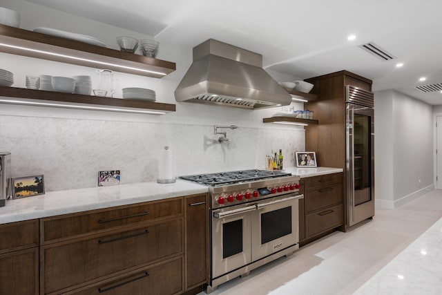 kitchen featuring high quality appliances, backsplash, wall chimney range hood, light stone countertops, and dark brown cabinetry