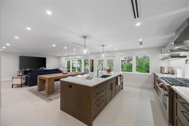 kitchen featuring a center island with sink, double oven range, sink, light stone counters, and wall chimney range hood