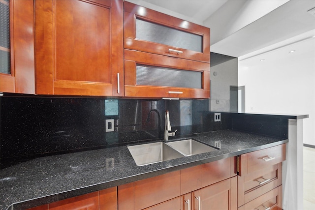 kitchen with decorative backsplash, glass insert cabinets, brown cabinets, dark stone countertops, and a sink