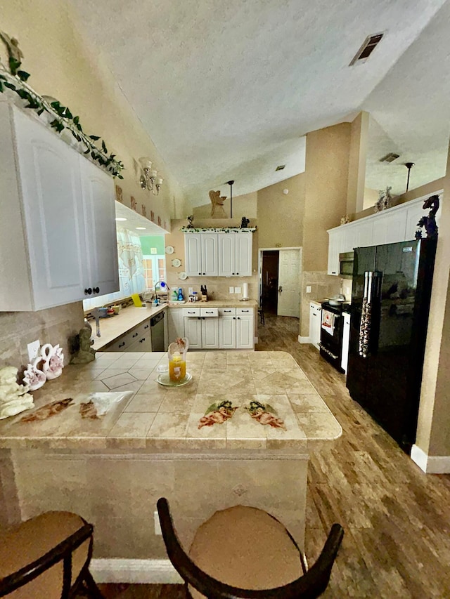 kitchen with kitchen peninsula, tile countertops, vaulted ceiling, white cabinets, and black appliances