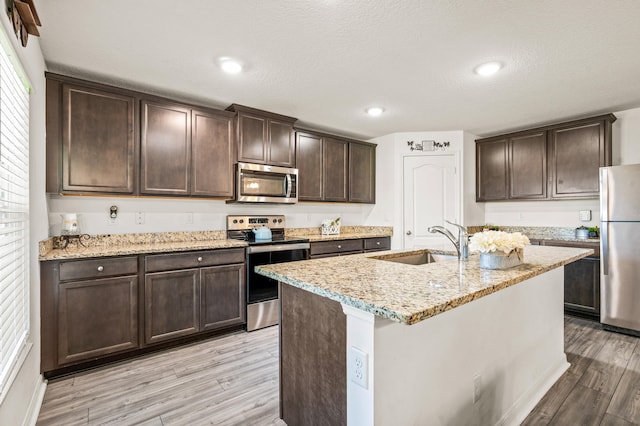 kitchen with light stone counters, sink, appliances with stainless steel finishes, a center island with sink, and light hardwood / wood-style floors