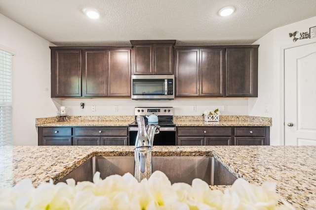 kitchen with a textured ceiling, stainless steel appliances, light stone countertops, and dark brown cabinetry