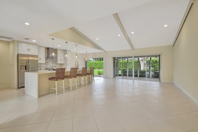 interior space with vaulted ceiling with beams, wall chimney range hood, hanging light fixtures, a center island with sink, and stainless steel fridge