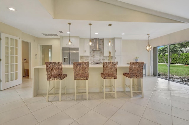 kitchen featuring wall chimney exhaust hood, stainless steel refrigerator with ice dispenser, a kitchen bar, pendant lighting, and white cabinetry