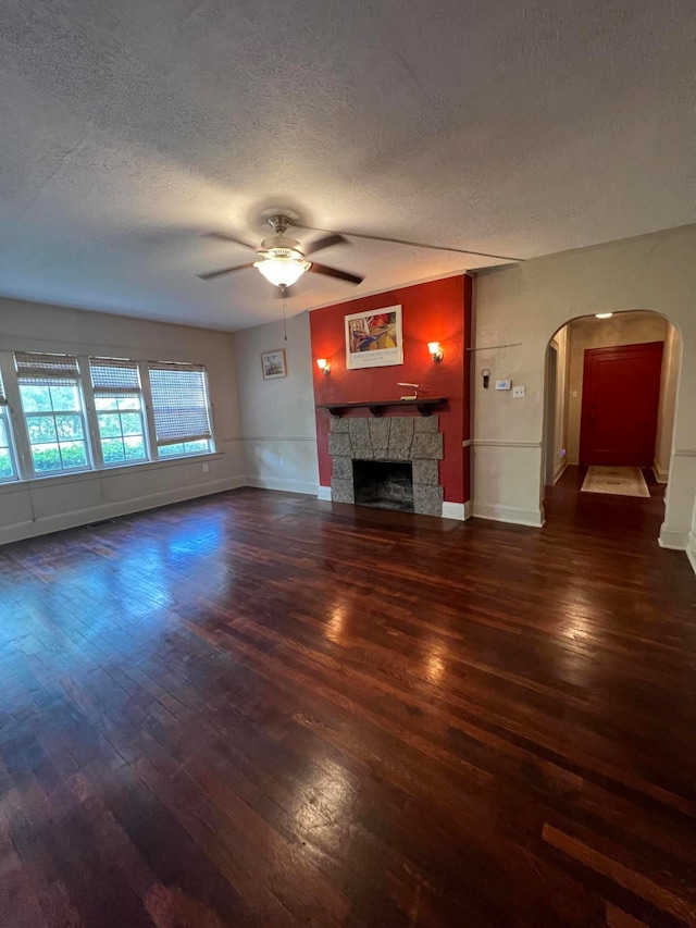 unfurnished living room with a textured ceiling, a stone fireplace, ceiling fan, and dark wood-type flooring