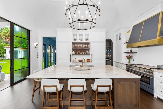 kitchen featuring an inviting chandelier, stainless steel stove, custom exhaust hood, a large island with sink, and white cabinetry