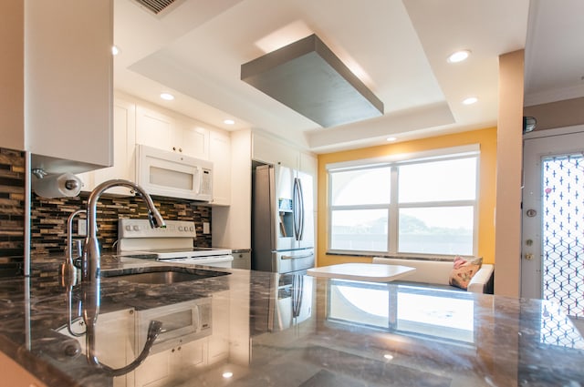 kitchen with a raised ceiling, white appliances, backsplash, white cabinetry, and dark stone counters