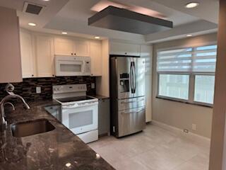 kitchen with a tray ceiling, white cabinets, a sink, dark stone counters, and white appliances