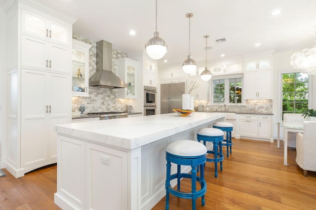 kitchen featuring wall chimney exhaust hood, a spacious island, stainless steel appliances, white cabinetry, and light wood-type flooring